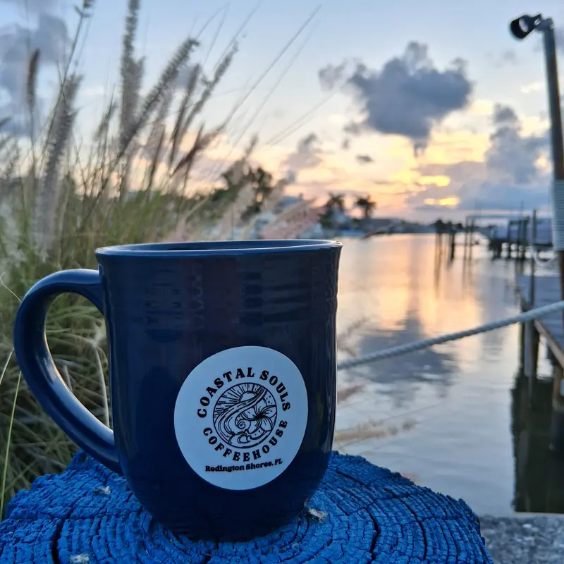 A navy Coastal Souls mug on a wooden post by the water, morning sky over the marina