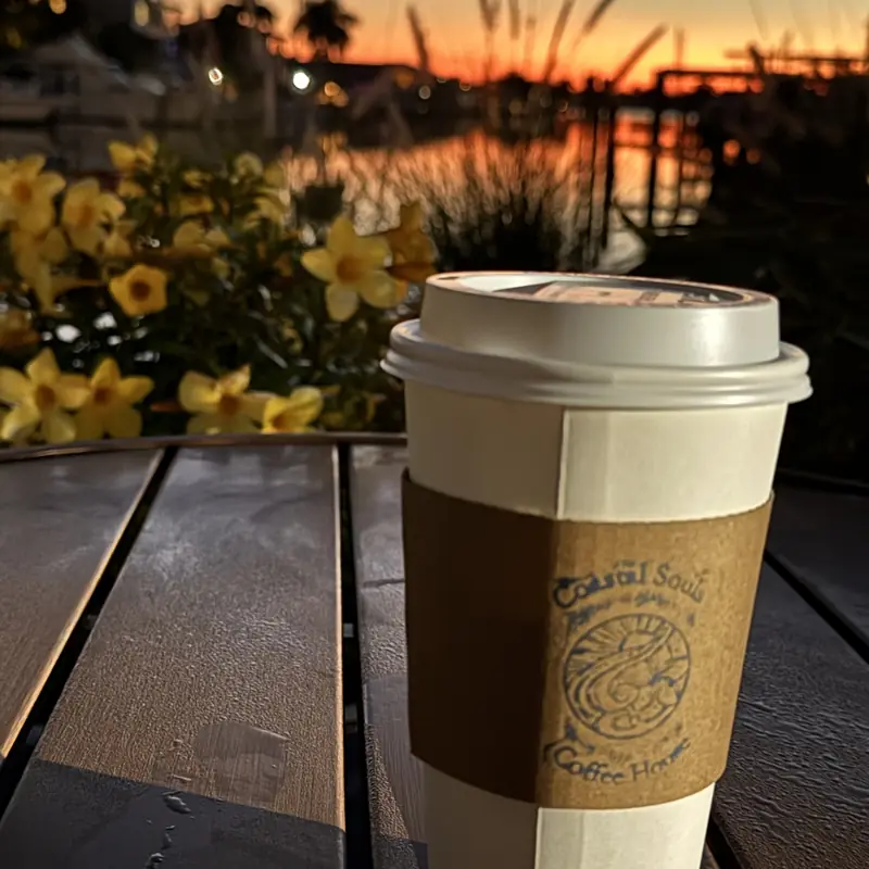A Coastal Souls cup on a patio table in the early morning, sky over the marina